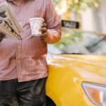 Crop unrecognizable ethnic male cab driver with plastic glass of beverage reading newspaper near contemporary car in town — I